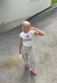 White T-shirt with red text and bow graphic, patterned grey snake print trousers, pink slide sandals, holding a clear cup, blonde hair.