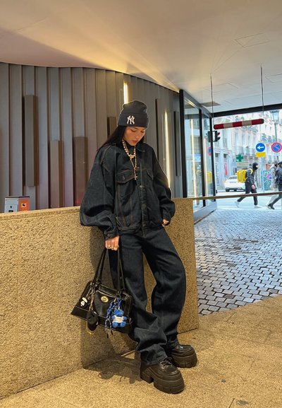 Chaqueta de mezclilla oscura de gran tamaño y pantalones, gorro negro, zapatos de plataforma gruesos y un bolso negro con accesorios de llavero coloridos.