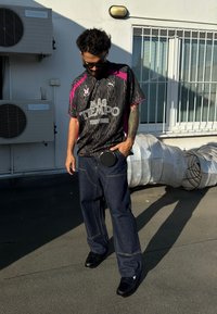 Zwart shirt met roze accenten, voorzien van een logo en tekst, gecombineerd met losse donkere spijkerbroeken met zichtbare stiksels en zijzakken.