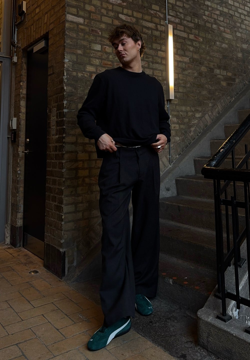 Svart langermet skjorte, løse svarte bukser og grønne sko på en strukturert murvegg, nær trapper og en døråpning.