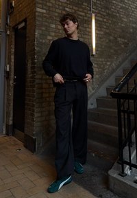 Svart langermet skjorte, løse svarte bukser og grønne sko på en strukturert murvegg, nær trapper og en døråpning.