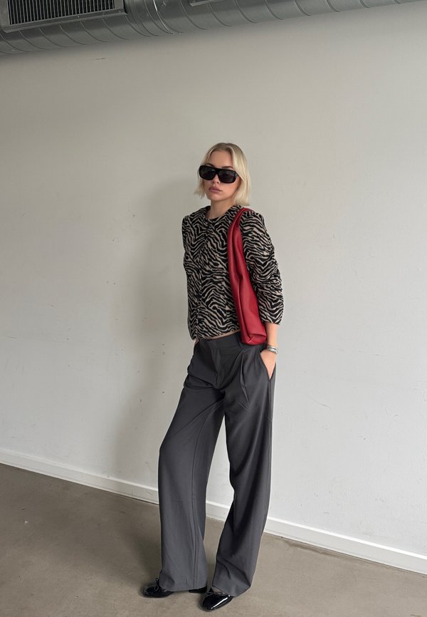 Une femme présente un haut à imprimé zèbre, un pantalon gris à jambes larges, des lunettes de soleil noires et un sac rouge porté à l’épaule, devant un mur uni.