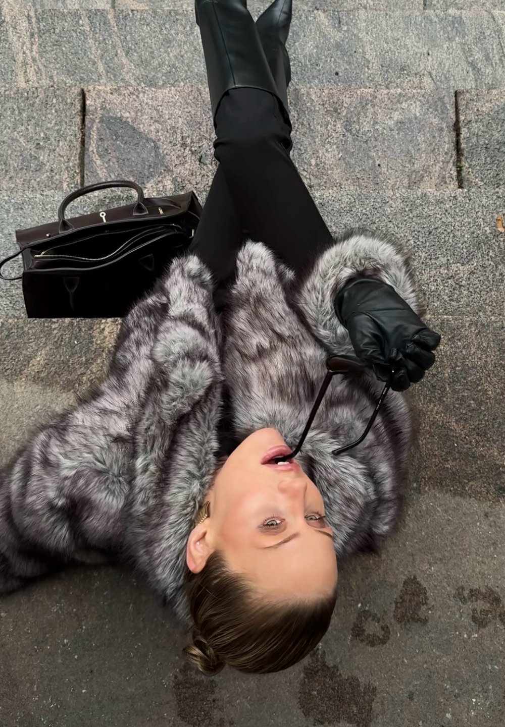 Szary płaszcz z sztucznego futra o miękkiej fakturze, czarne skórzane kozaki sięgające kolan, czarna torebka i okulary trzymane w ustach, umieszczone na kamiennej powierzchni.