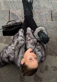 Szary płaszcz z sztucznego futra o miękkiej fakturze, czarne skórzane kozaki sięgające kolan, czarna torebka i okulary trzymane w ustach, umieszczone na kamiennej powierzchni.