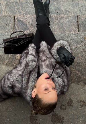 Abrigo gris de piel sintética con textura suave, botas negras de cuero hasta la rodilla, bolso negro y gafas sujetas en la boca, colocados sobre una superficie de piedra.