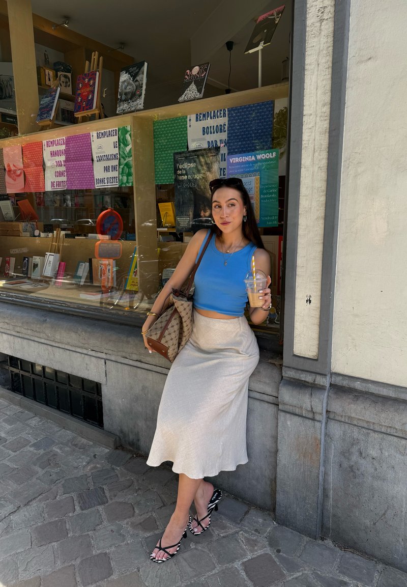 Femme en haut bleu et jupe blanche s'appuie contre la vitrine d'une librairie en tenant un café glacé, avec un sac à main à motifs sur l'épaule.