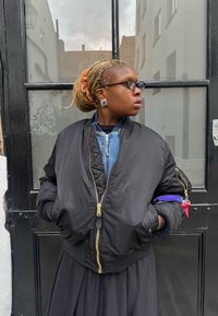 Svart bomberjakke med gullglidelås, brukt over en denimtopp. Lang svart skjørt. Sølvfargede kvadratiske øredobber, oransje hårstrikk, briller.