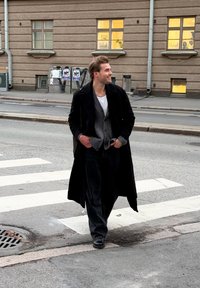 Crni dugi kaput preko sive veste, bijele majice i tamnih hlača. Stoji na pješačkom prijelazu s nogostupom i zgradama u pozadini.