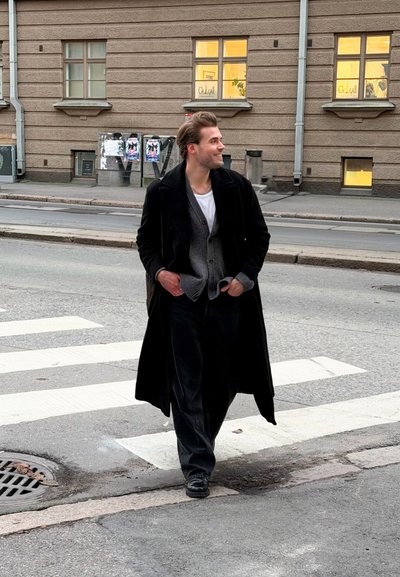 Abrigo largo negro sobre un cárdigan gris, camiseta blanca y pantalones oscuros. De pie en un paso de peatones con la acera y edificios al fondo.