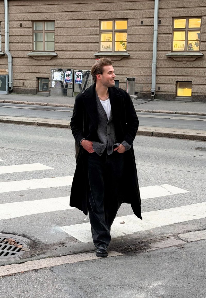 Cappotto nero lungo sopra un cardigan grigio, maglietta bianca e pantaloni scuri. In piedi su un attraversamento pedonale con marciapiede e edifici sullo sfondo.