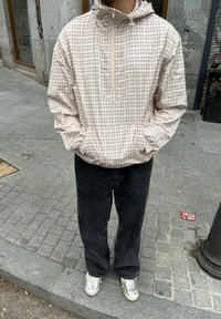 Light brown and cream checkered pullover jacket with a half-zip, front pocket, and adjustable hood, teamed with black wide-leg trousers and silver trainers.