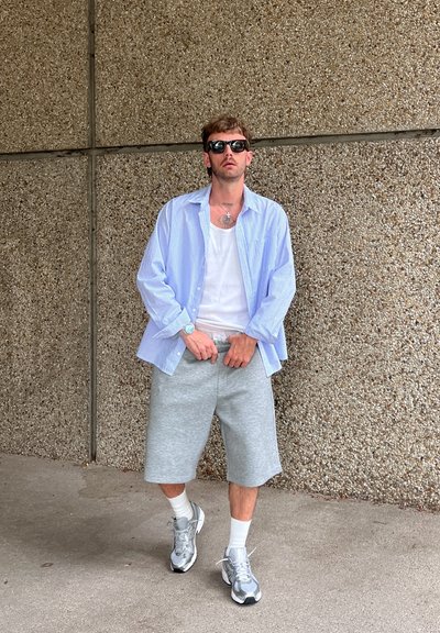 Camisa de botones a rayas azul claro sobre una camiseta blanca sin mangas, combinada con pantalones cortos grises y zapatillas plateadas. Los accesorios incluyen gafas de sol y un reloj.