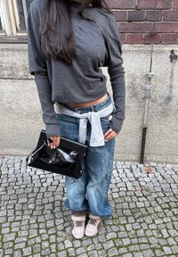 Top gri cu mâneci lungi, talie legată deschisă la culoare peste blugi largi albaștri, poșetă lucioasă neagră și ghete maro deschis pe o suprafață cu piatră cubică.