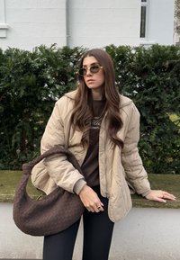 Μπεζ ραφτό μπουφάν με φερμουάρ, καφέ πλεκτό πουλόβερ, μαύρες κολάν και ένα σκούρο καφέ υφασμάτινο τσάντα με καμπύλη λαβή. Φόντο με πράσινα θάμνους.