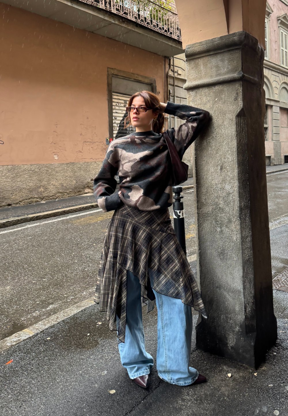 Strikket camouflage-trøje, ternet asymmetrisk nederdel, blå jeans med vide ben, brune spidse sko og mørk skuldertaske i bymiljø.