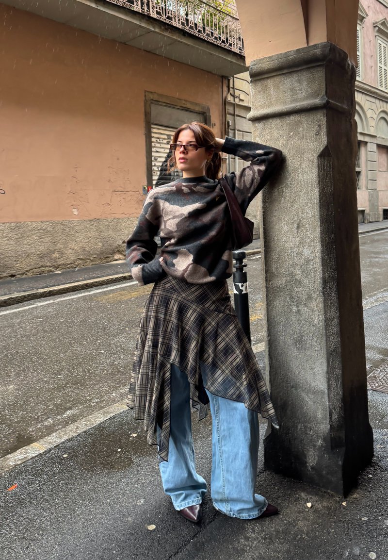 Camisola de malha camuflada, saia assimétrica às riscas, jeans azul de perna larga, sapatos marrons com bico fino e mala de ombro escura em ambiente urbano.