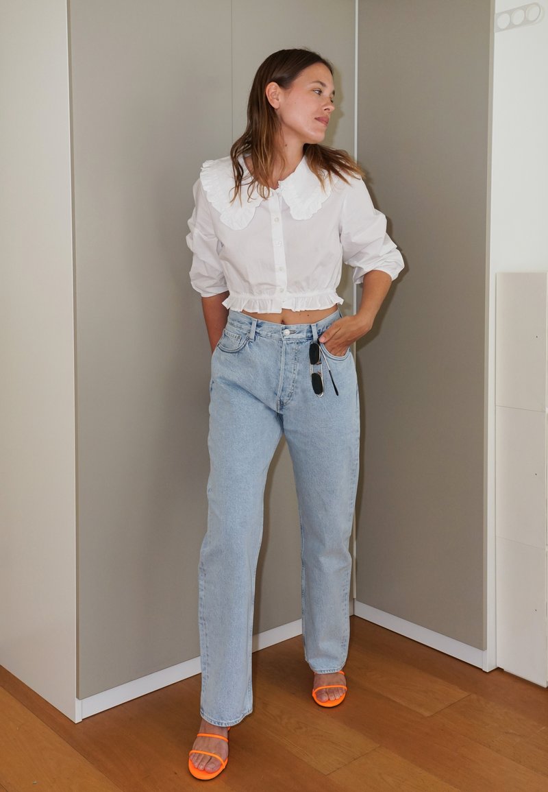 Valkoinen lyhythihainen paita, jossa on röyhelöhelma ja suuri kaulus, yhdistettynä vaaleansinisiin suoriin farkkuihin ja oransseihin korkokenkiin.