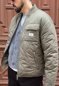 Giacca bomber trapuntata verde oliva con zip e dettagli con tasche. Il materiale appare morbido, leggero e idrorepellente con una texture liscia.