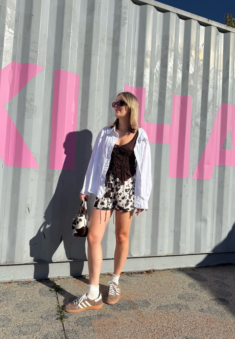 Chemise blanche oversize, top marron froncé, shorts à imprimé vache noir et blanc. Baskets marron avec accents blancs, sac à main assorti et chaussettes blanches.