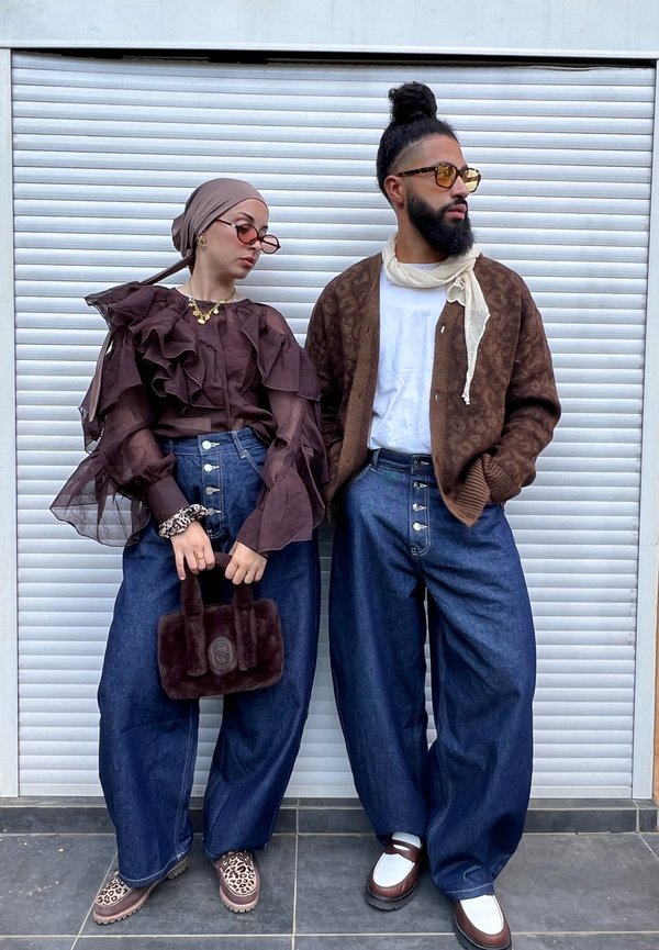 Een paar individuen die laagjes bruin gerimpelde tops dragen, brede blauwe denimbroeken en unieke accessoires zoals een harige tas en stijlvolle schoenen.