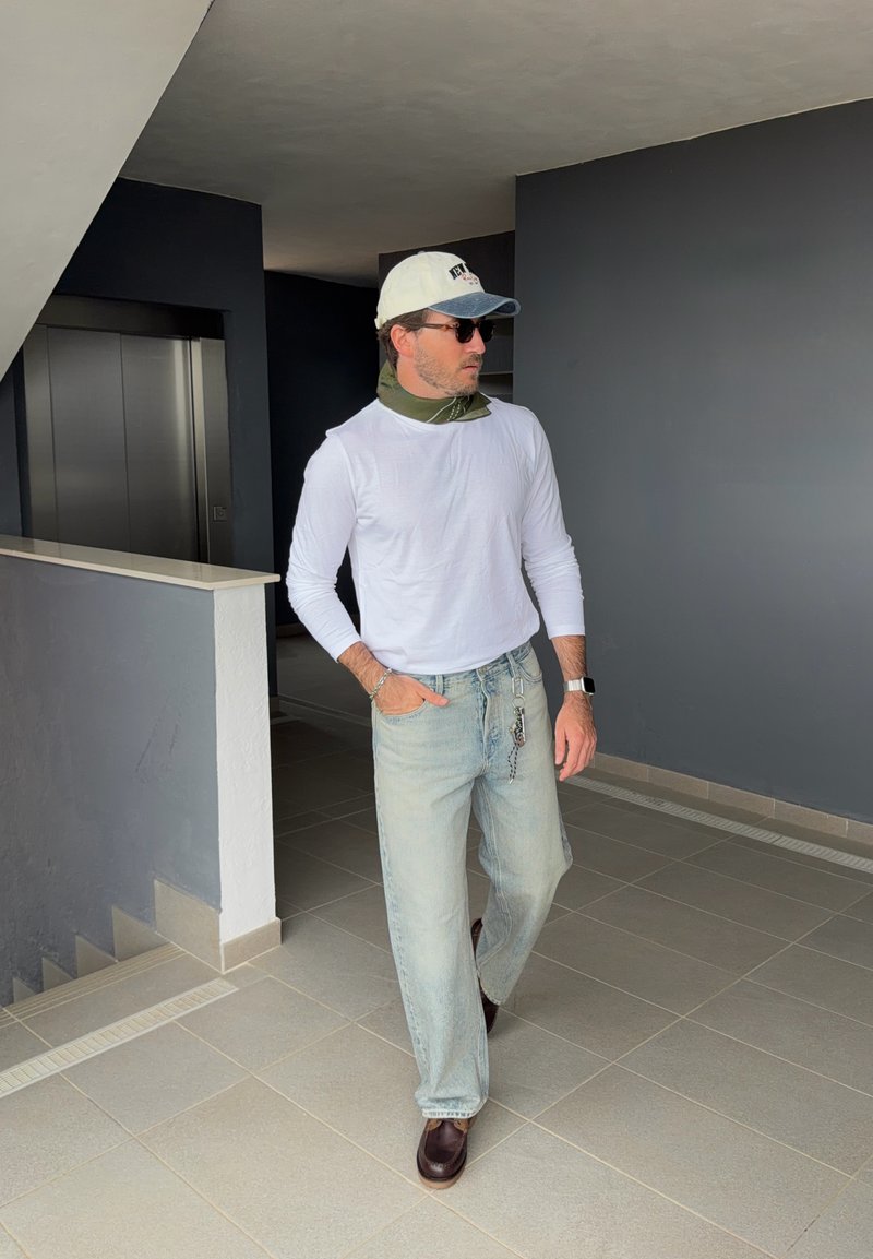Homme portant une chemise blanche à manches longues, un jean bleu clair, une casquette blanche, des lunettes de soleil et des chaussures marron, marchant dans un couloir intérieur moderne près de l'ascenseur.