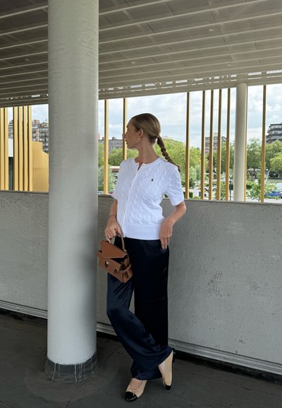 Cárdigan blanco de punto con manga corta, combinado con pantalones de satén azul marino y zapatos planos beige. Sosteniendo un bolso estructurado marrón.