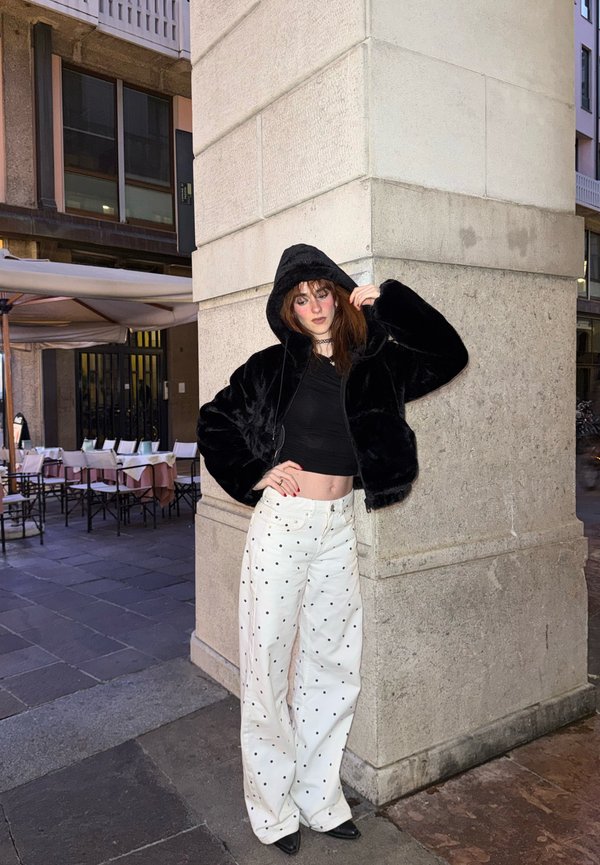 Donna con giacca nera con cappuccio e top corto nero, pantaloni bianchi a pois, appoggiata a un pilastro di pietra in una strada cittadina di notte.