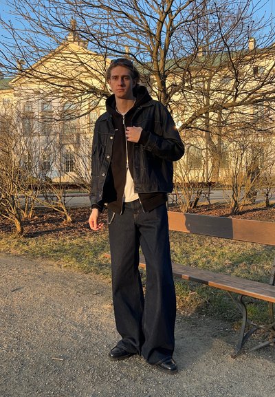 Chaqueta de mezclilla oscura sobre un hoodie negro, combinada con jeans negros de pierna ancha y zapatos negros. El fondo presenta árboles sin hojas y un edificio.