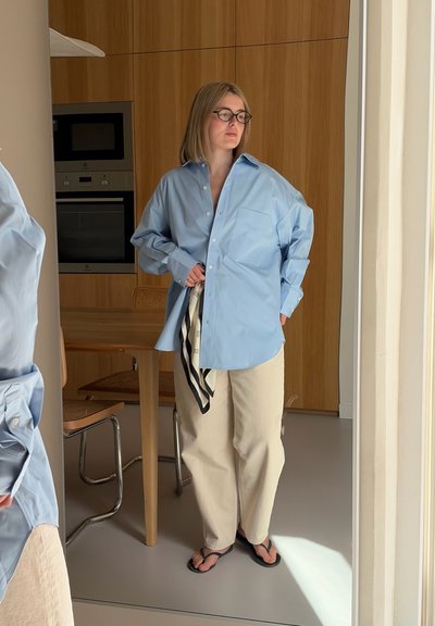 Mujer con gafas, camisa azul clara oversize, pantalones beige y chanclas negras, está de pie en el interior cerca de una mesa de madera y electrodomésticos de cocina.