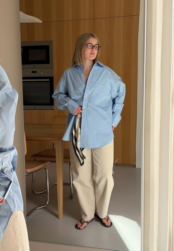 Femme portant des lunettes, une chemise bleu clair oversize, un pantalon beige et des tongs noires, debout à l'intérieur près d'une table en bois et d'appareils de cuisine.