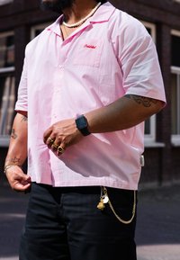 Camisa de manga curta rosa claro com um bolso no peito apresentando "Proibido" em costura vermelha. Calças pretas com uma corrente dourada e charms.