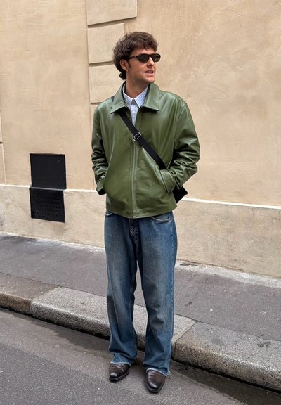 Chaqueta de cuero verde, camisa a cuadros azul claro, jeans azules holgados, zapatos negros y gafas de sol; de pie en una calle de la ciudad con una pared texturizada.