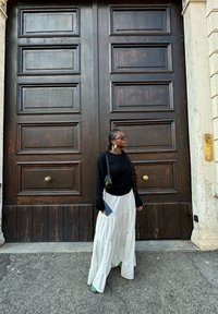 Maglione nero a maniche lunghe, gonna maxi bianca a strati, scarpe verdi. Capelli intrecciati, in piedi davanti a grandi porte di legno con hardware in ottone.