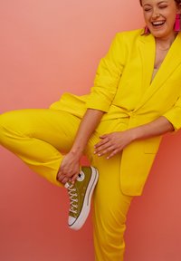 Abito giallo brillante realizzato in tessuto liscio, con blazer aderente e pantaloni a sigaretta. Sneakers in tela verde con dettagli bianchi. Sfondo rosa.