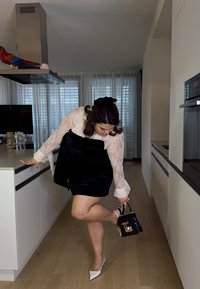 Vestido preto de veludo sobre uma blusa branca de renda, combinado com sapatos de salto brancos pontiagudos e uma pequena bolsa preta com detalhes em dourado.