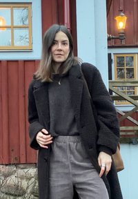 Manteau noir texturé sur un pull sombre, associé à un pantalon en velours côtelé gris clair. Arrière-plan naturel avec un bâtiment en bois rouge et des fenêtres.