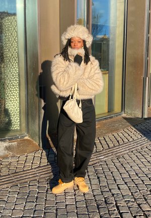 Puhast bež plašč z usklajeno krzna klobukom, črnimi hlačami in čevlji v karamelni barvi. Drži svetlo bež torbico na rami. Odtis iz cobblestone.