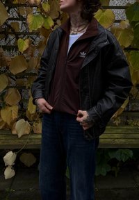 Sort læderjakke over en brun lynlås-sweatshirt, parret med blå jeans. Teksturerne kontrasterer mellem materialerne; glat og afslappet stil.