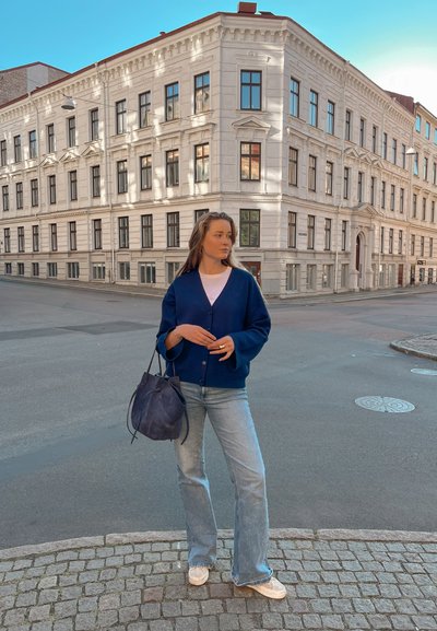 Cárdigan azul marino con un escote en V profundo y botones, combinado con jeans acampanados de mezclilla clara. Lleva un bolso tipo cubo oscuro. Fondo de calle de adoquines.
