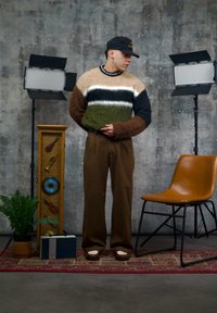 Pull en tricot à rayures vertes, marron, noires et blanches, porté avec un pantalon marron. Comprend une casquette noire et des chaussures à semelle blanche.