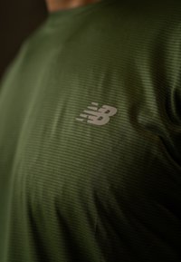 Olivengrønn trenings-T-skjorte med teksturert stoff og en lys grå New Balance-logo på venstre bryst.