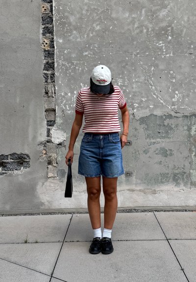 Camisa de rayas de manga corta en rojo y blanco, combinada con pantalones cortos de mezclilla. Lleva zapatos negros y una gorra de béisbol blanca, sosteniendo una bolsa negra.