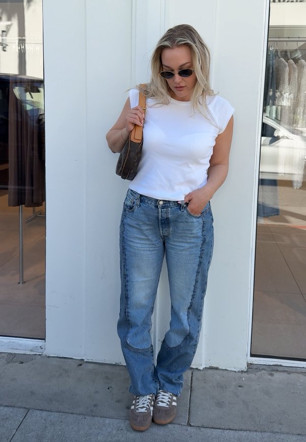 Femme portant un débardeur blanc, un jean bleu, des baskets et des lunettes de soleil, tenant un sac à main marron, debout contre un mur blanc près d'une vitrine de magasin.