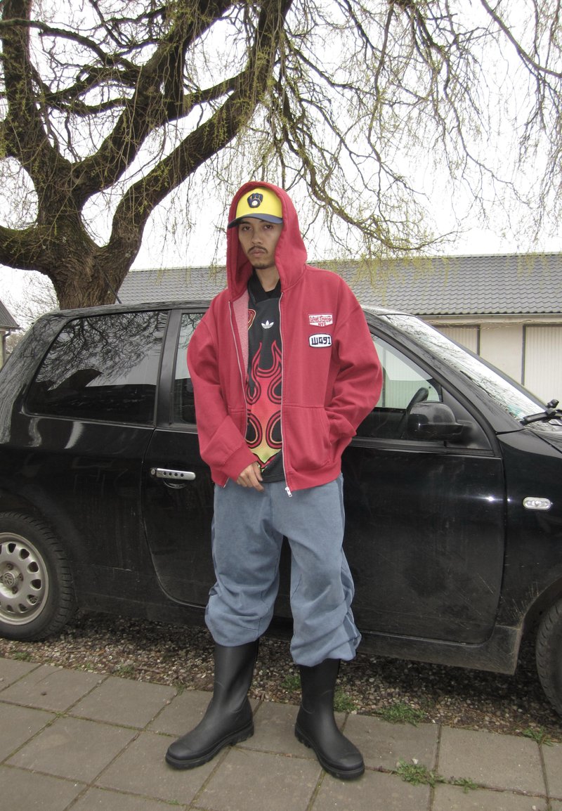 Jeune homme en sweat à capuche rouge et chemise à motif de flammes se tient les mains dans les poches à côté d'une voiture noire sous un arbre sans feuilles.