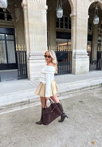 Off-white skulderfri genser, beige plissert miniskjørt, brune knelange støvler og en mørk brun semsket skinnveske. Stensøyler i bakgrunnen.