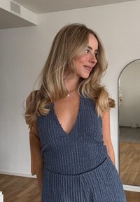 Blå texturerad klänning med djup V-ringning och elastisk midja. Bärs med en ljusbrun handväska, mot en neutral bakgrund med en spegel.