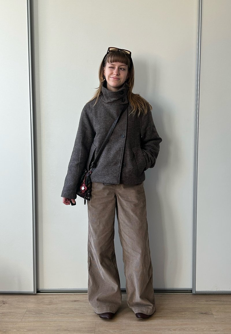 Jeune femme debout à l'intérieur portant une veste gris foncé, un pantalon beige à jambes larges, des lunettes de soleil sur la tête et un petit sac bandoulière, faisant un clin d'œil.