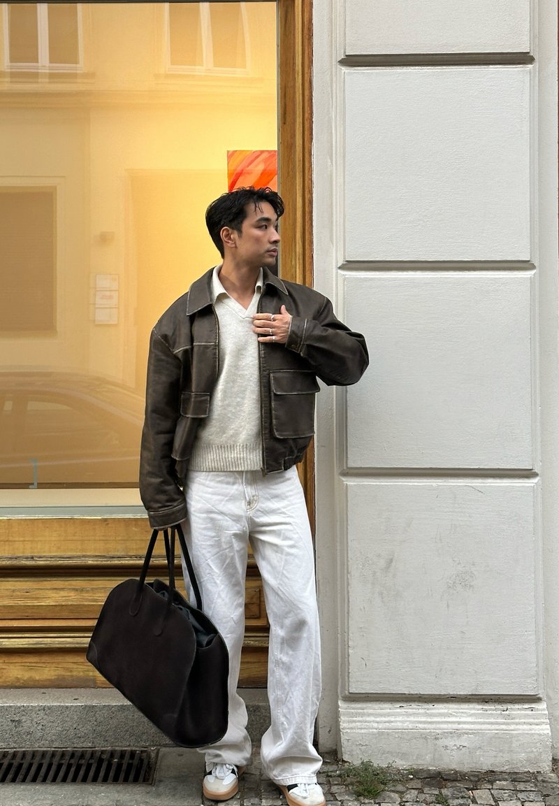 Καφέ δερμάτινο jacket πάνω από ένα ανοιχτόχρωμο πουλόβερ, λευκά παντελόνια σε φαρδιά γραμμή και λευκά αθλητικά παπούτσια. Κρατάει μια μαύρη τσάντα tote. Ουδέτερο εξωτερικό φόντο.