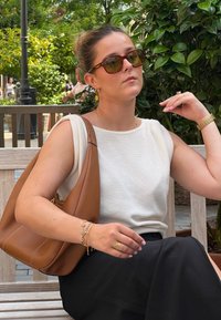 Ανοιχτό μπεζ αμάνικο τοπ, μαύρη φούστα, μεγάλη καφέ τσάντα. Αξεσουάρ περιλαμβάνουν χρυσά σκουλαρίκια, βραχιόλι και δαχτυλίδι. Ο ήλιος φιλτράρεται μέσα από την πράσινη βλάστηση.