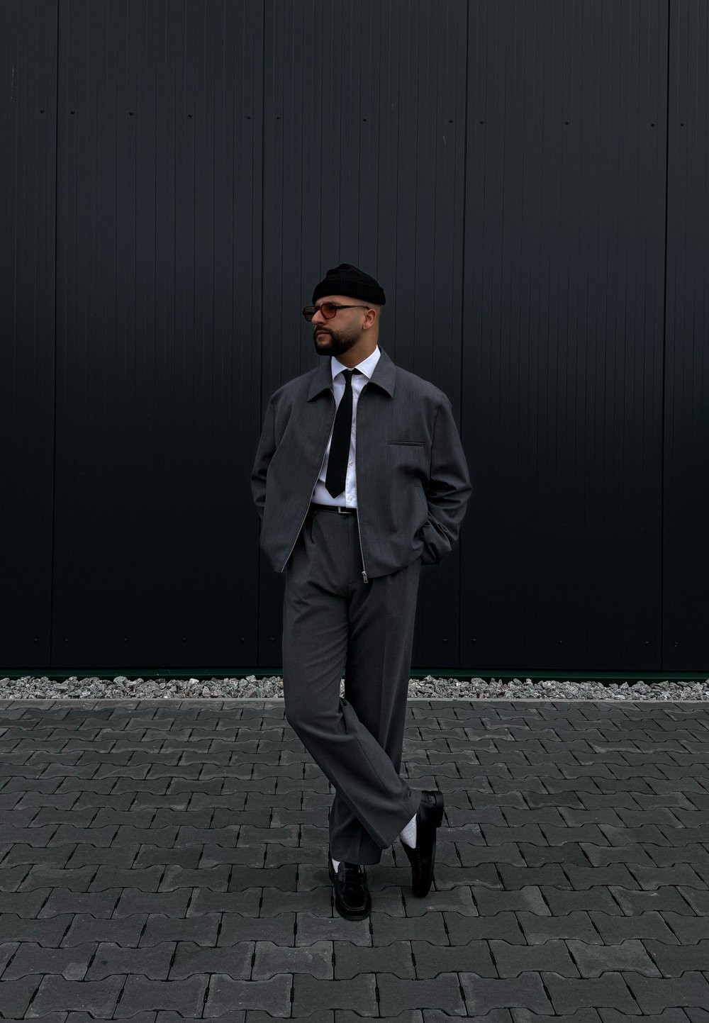 Γκρίζα ζακέτα με φερμουάρ πάνω από άσπρο πουκάμισο και μαύρη γραβάτα, συνδυασμένη με γκρίζα παντελόνια και μαύρα παπούτσια. Ο φόντος περιλαμβάνει ένα σκοτεινό τοίχο και pave.